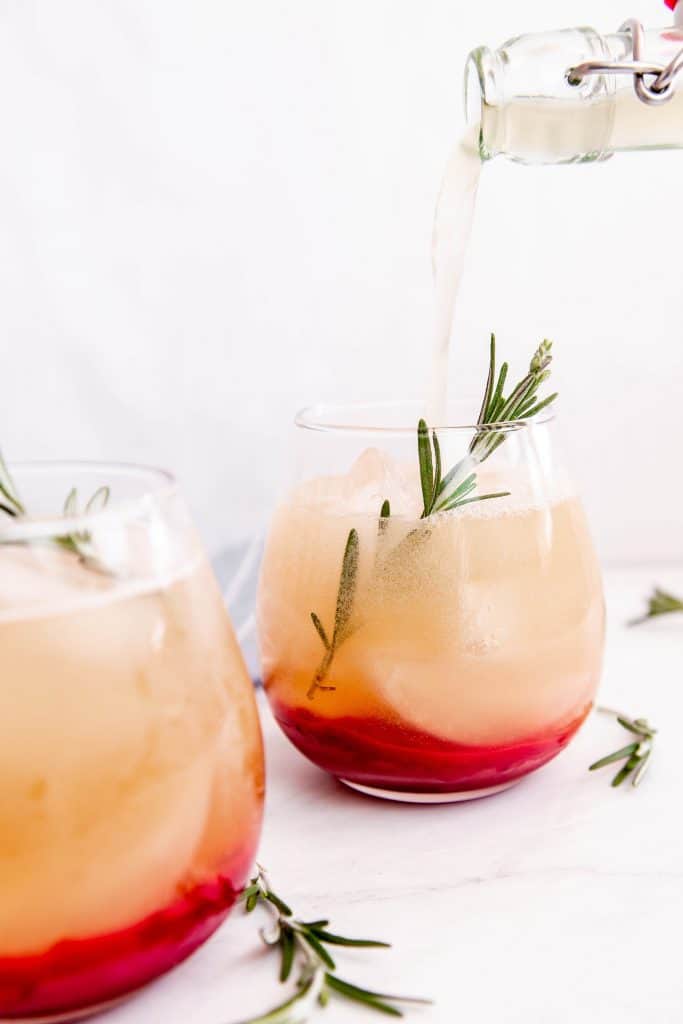 a glass of cranberry ginger kombucha being filled. A sprig of rosemary sticking out of the glass.