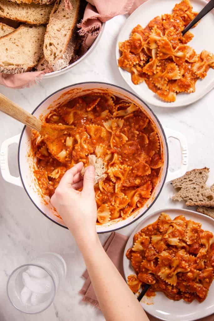 hand dipping a piece of sourdough bread into a dutch oven full of vegan chickpea bolognese.