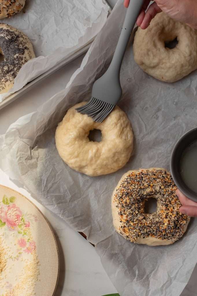 Brushing unbaked sourdough bagel with an egg white egg wash before adding toppings.