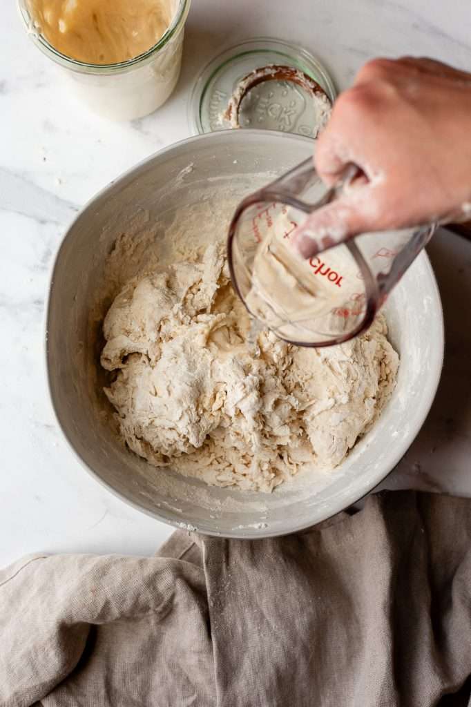 Person adding water to sourdough bagel dough in a mixing bowl.