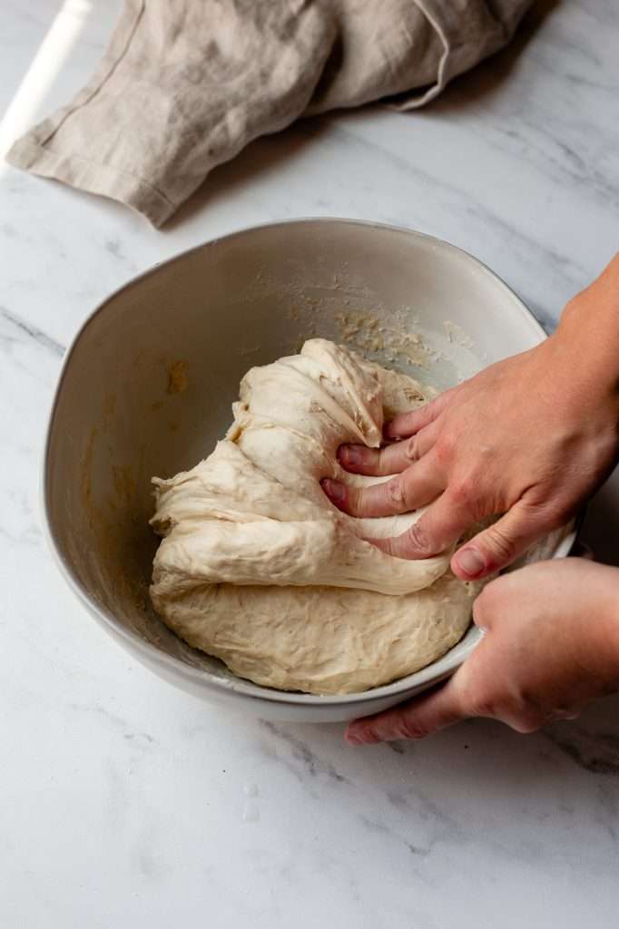 Person folding sourdough bagel dough back into the bowl after stretching.