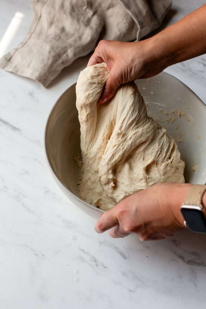 Person stretching sourdough bagel dough upwards from a mixing bowl.