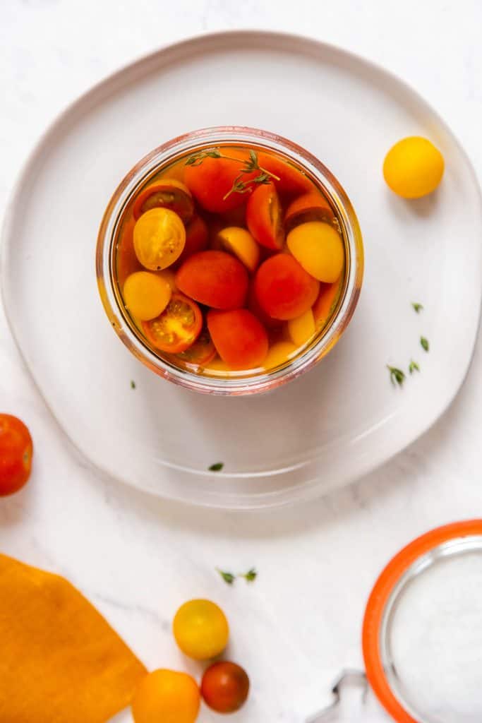 Orange, red and yellow cherry tomatoes, sliced in half in a jar with brine. The jar sits on a white plate. 