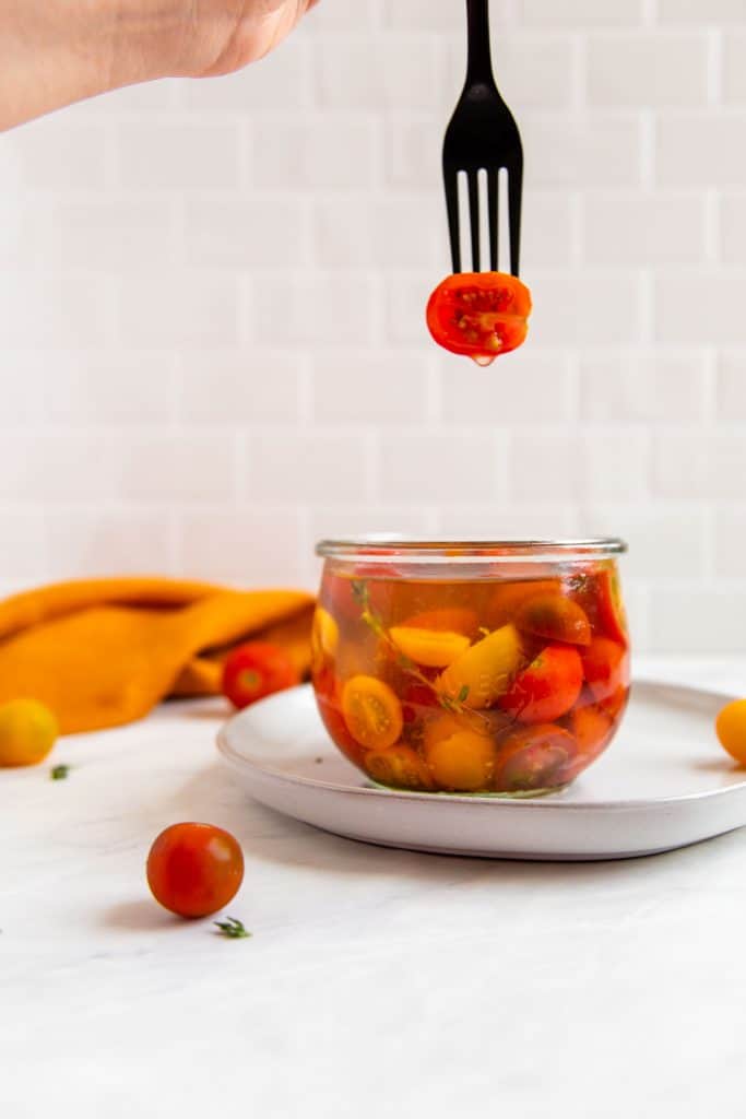 Yellow, orange and red fermented cherry tomatoes in a jar with brine. One cherry tomatoes is speared with a fork. 