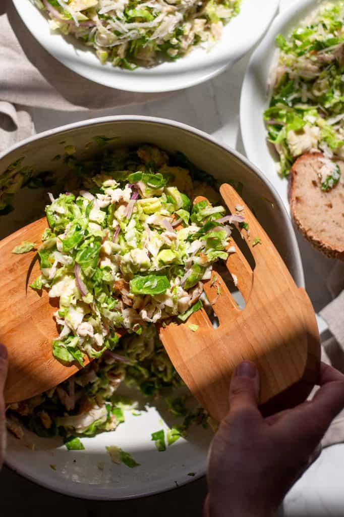 someone uses salad scoops to serve Warm Brussel Sprout Caesar Salad with Chicken and Parmesan into bowls. 