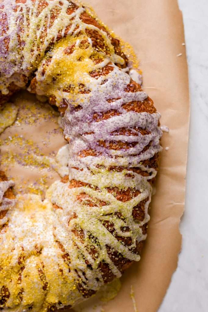 sourdough king cake on a piece of brown parchment paper, decorated with icing and purple, gold and green sprinkles. 