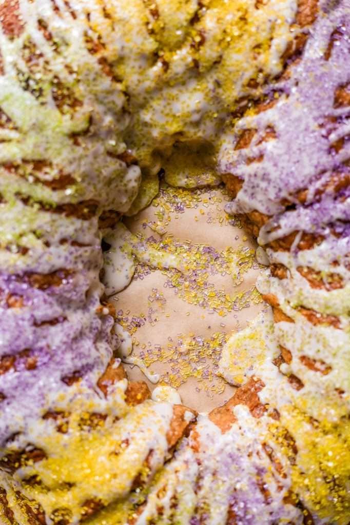 A close-up of a freshly baked sourdough king cake, drizzled with white icing and topped with vibrant purple, green, and gold sugar sprinkles.