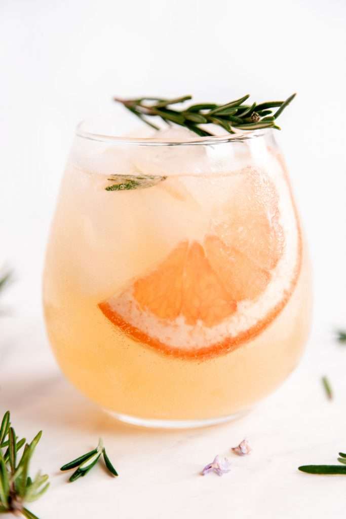 a slice of grapefruit floats in a stemless wine glass filled with grapefruit and rosemary flavored kombucha. A sprig of rosemary lays atop the glass.