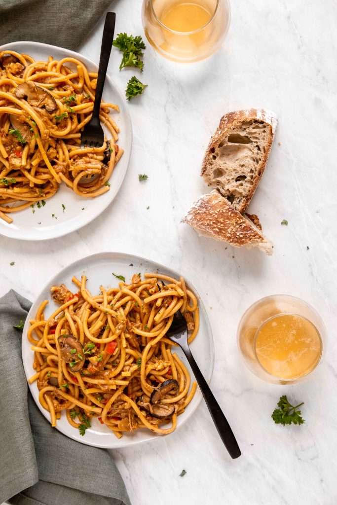 two plates of cajun veggie pasta on a white marble surface. A side of crusty sourdough bread sits next to the plates. 