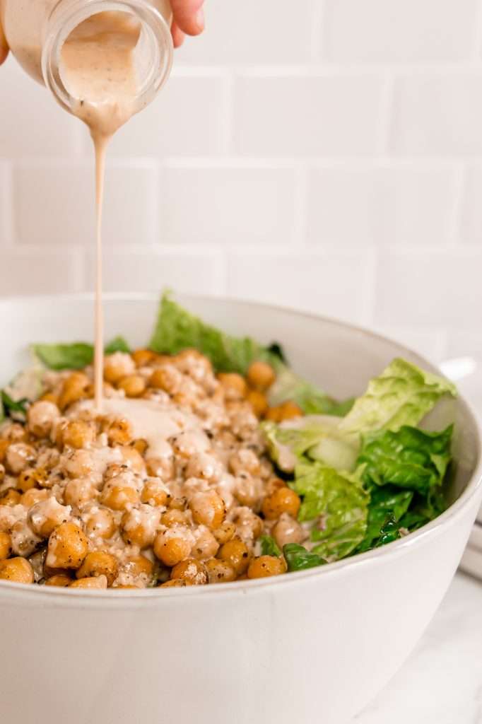 someone pouring ceasar dressing over romaine lettuce and roasted chickpeas in a big white salad mixing bowl. 