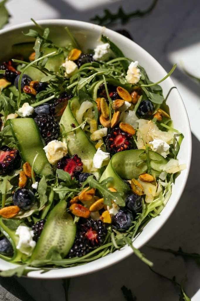 sauerkraut and arugula salad in a white bowl with goat cheese, blackberries, long sliced cucumbers, and pistachios on top. Honey mustard dressing is drizzled over the salad. 