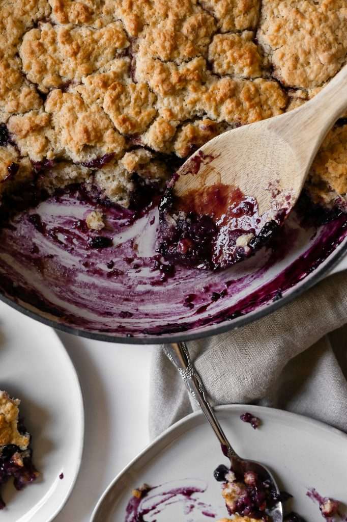 blackberry dutch oven cobbler cooked in a white dutch oven, part of the cobbler has been scooped out and served with a wooden spoon.