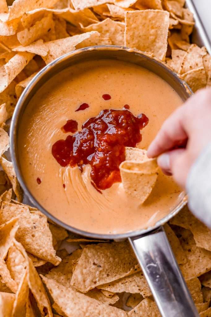 cottage cheese queso melted in a small pot, topped with salsa. The pot of queso is surrounded by tortilla chips and someone is dipping a chip into the queso. 