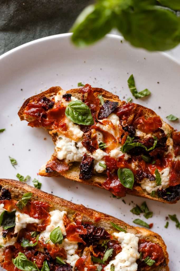 pizza cottage cheese toast topped with crispy prosciutto and sun dried tomatoes. The toast has a bite taken out of it and is resting on a white plate. 