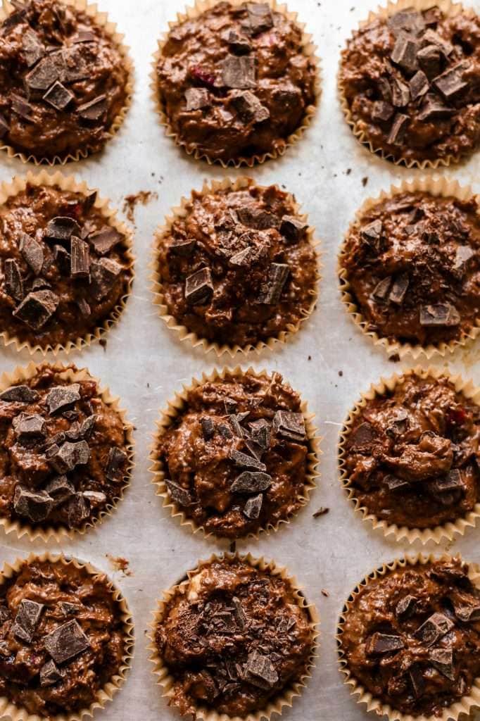 chocolate cherry sourdough muffin batter spooned into a muffin pan lined with brown muffin liners. the muffins are topped with some tiny chocolate chunks. 