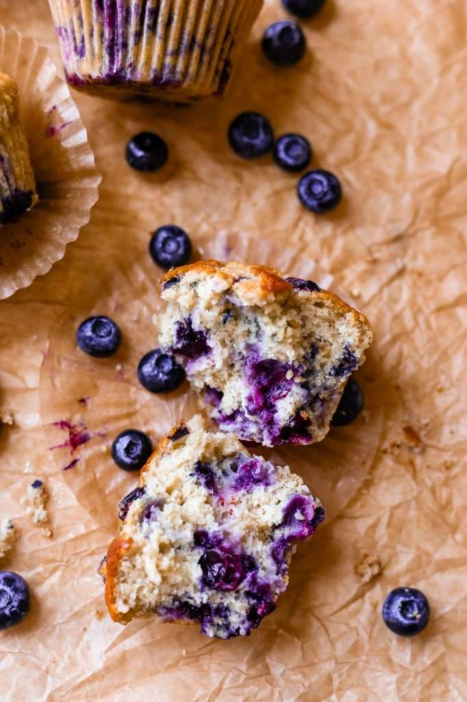 blueberry protein muffins on some crinkly brown parchment paper. One muffin is torn in half to display the moist and fluffy inner texture. 