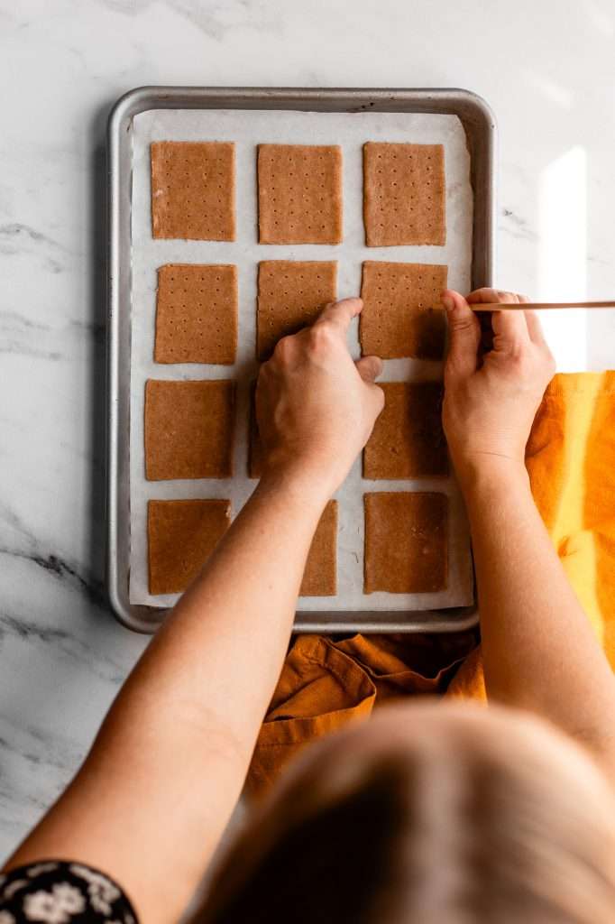 someone using a wooden kebab skewer to poke holes in the sourdough graham cracker dough. The dough has been cut into 2x2.5 inch rectangles and placed on a sheet pan. 