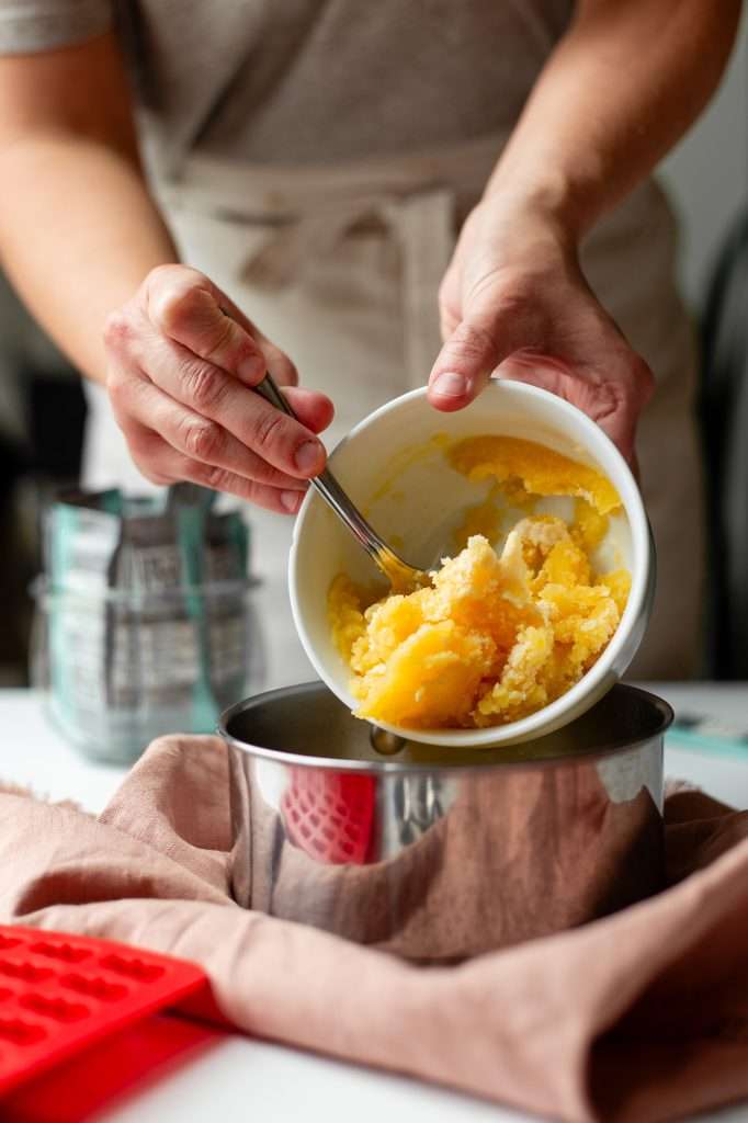 the third step to making protein gummies— Someone is scooping gelatin combined with cold fruit juice into a small silver pot of heated juice. 