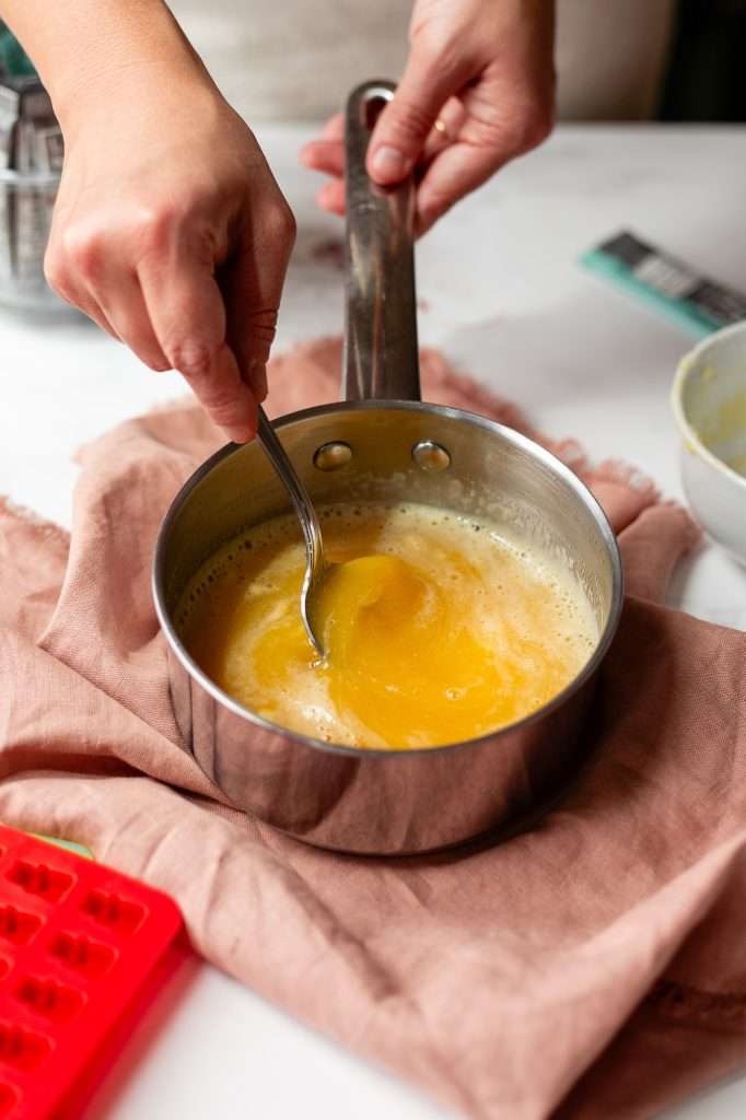 the third step to making protein gummies— Someone is using a spoon to mix the cold juice and gelatin mixture into the hot juice until evenly combined.  