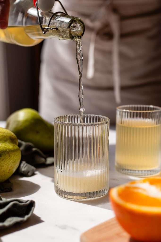 Pouring handmade perry, or pear cider, into a glass with fresh pears and an orange nearby.