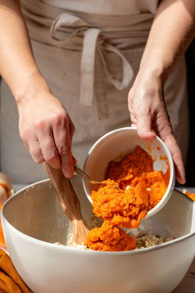 someone scooping pumpkin puree into the pumpkin protein muffins batter, to gently fold it in, without over-mixing. 