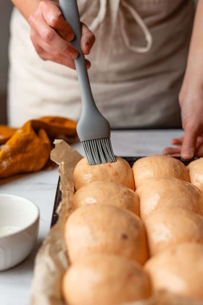 someone using a silicone pastry brush to brush the sourdough sweet potato dinner rolls with an egg white egg wash. 