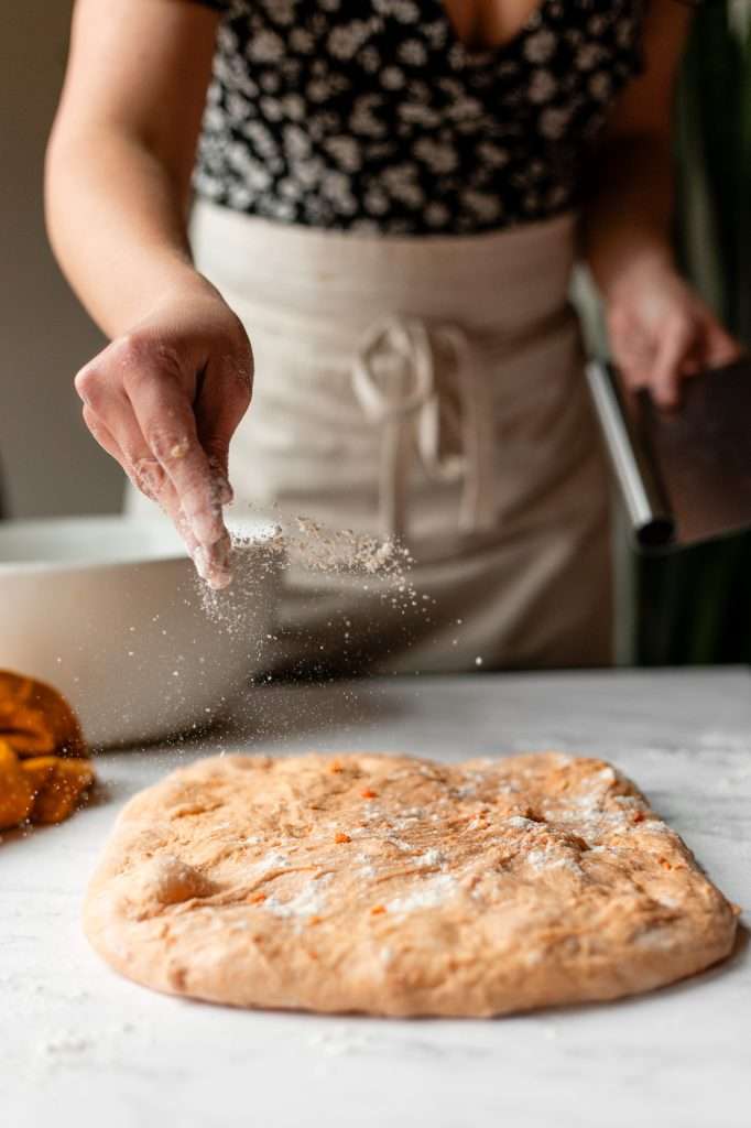 someone sprinkling flour on the sweet potato dinner roll dough that has been gently stretched out into a rectangle on a floured countertop. 