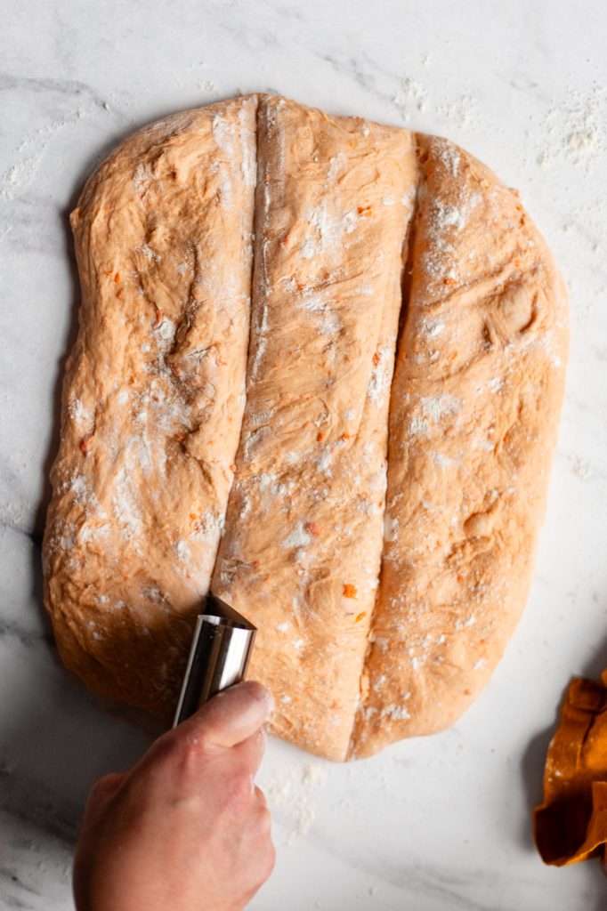 someone using a metal baking bench scraper to cut the sweet potato dinner roll dough into 12 even squares. 