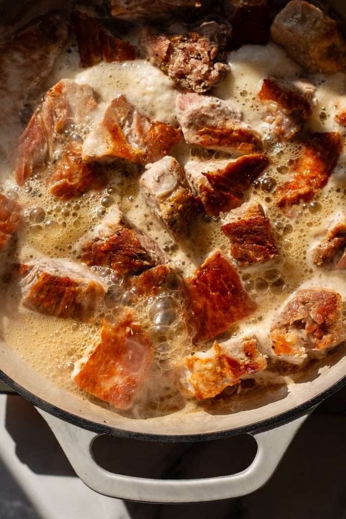 Pork loin chunks simmering in cider, with bubbling liquid forming around the browned pieces in a white Dutch oven.