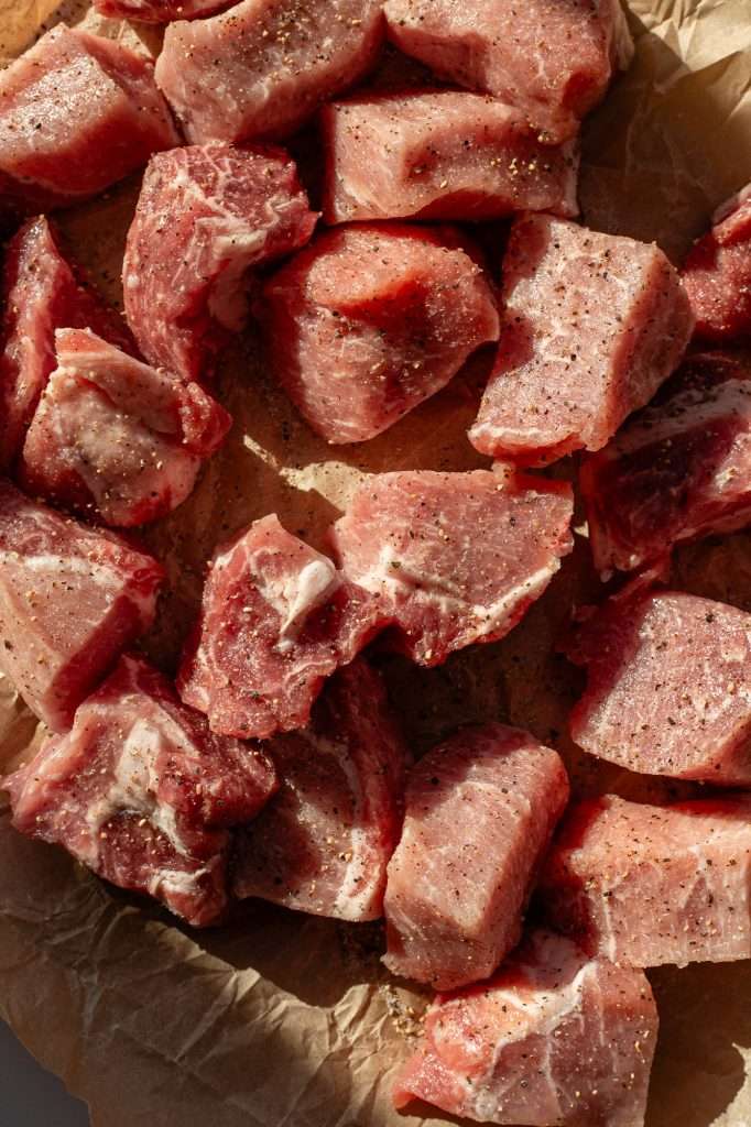 Raw pork loin chunks seasoned with salt and pepper, arranged on parchment paper in natural light.