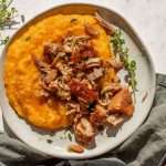 A plate of cider-braised pork and sauerkraut served over creamy butternut squash grits, garnished with fresh thyme and black pepper. The dish is photographed in natural light, with a rustic green napkin and fresh herbs in the background.