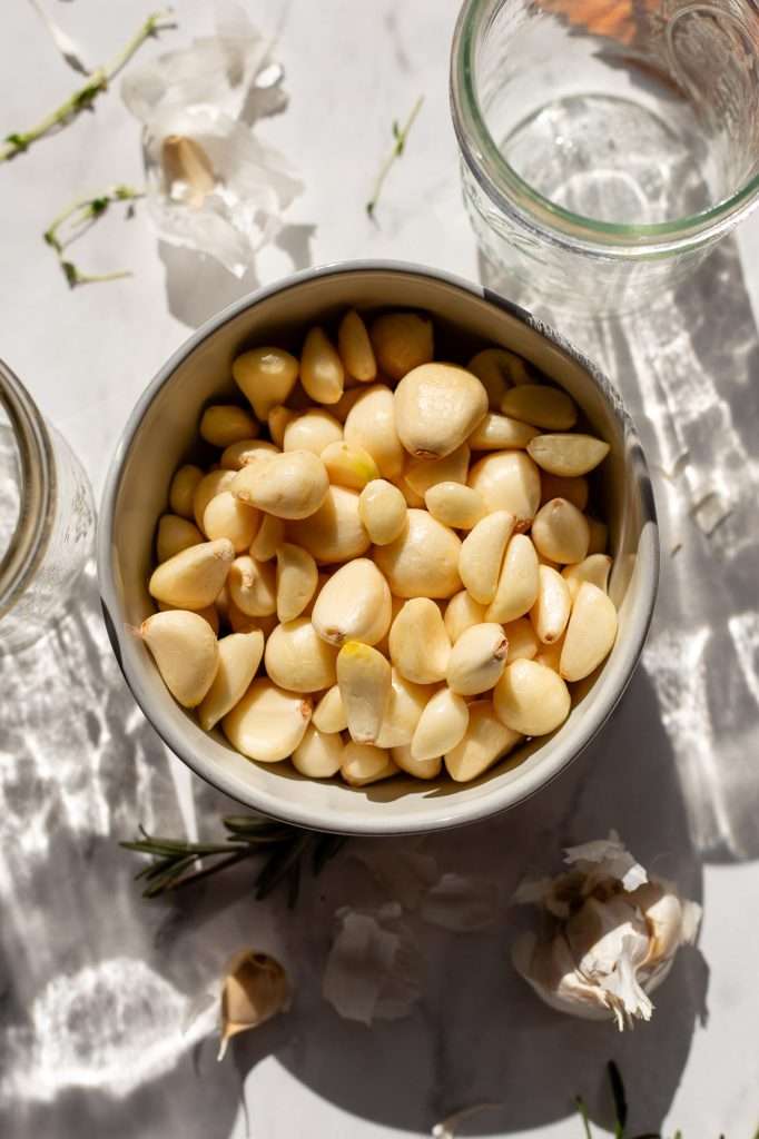 Bowl of peeled garlic cloves on a white marble countertop surrounded by empty jars, garlic skins, rosemary, and thyme sprigs.