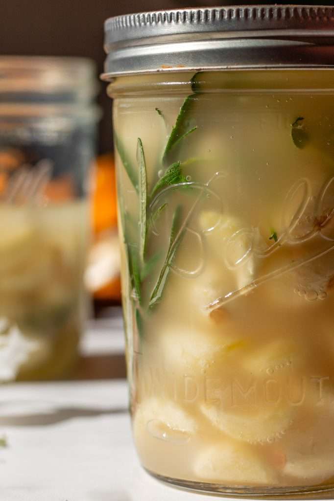 Close-up of a mason jar filled with probiotic pickled garlic cloves, fresh herbs like rosemary, and a tangy brine made with apple cider vinegar and sauerkraut brine. A second jar and garlic cloves are blurred in the background.