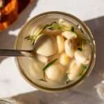 Overhead view of a jar filled with probiotic pickled garlic cloves, fresh herbs like thyme and rosemary, and homemade sauerkraut brine, with a spoon resting inside. Fresh garlic and a linen cloth are visible in the background.