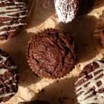 Wide view of sourdough gingerbread muffins on crinkled parchment paper, featuring muffins with drizzled eggnog icing, powdered sugar, and one muffin cut open to reveal its moist interior.