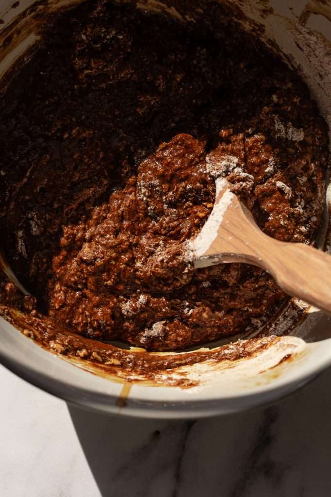 Thick and spiced sourdough gingerbread muffin batter being mixed with a wooden spoon in a white mixing bowl on a marble countertop.