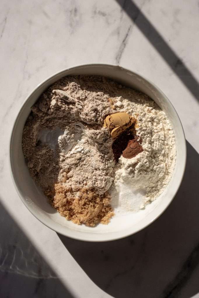 Overhead view of a mixing bowl with dry ingredients for sourdough gingerbread muffins, including whole wheat flour, all-purpose flour, brown sugar, baking soda, and spices like ginger and cinnamon.