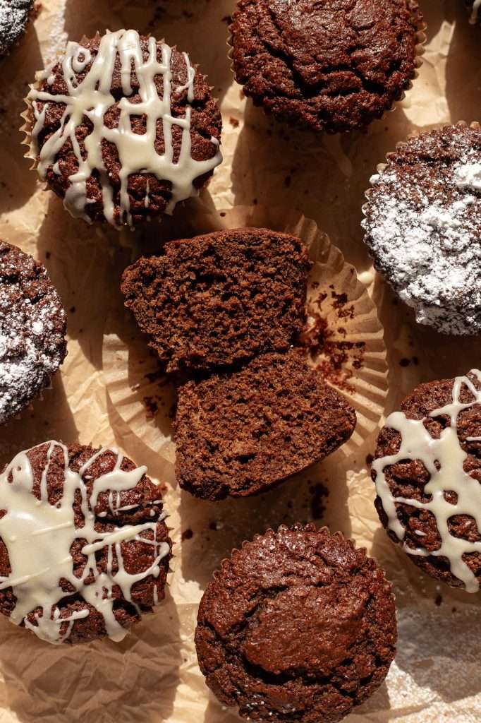 Close-up of sourdough gingerbread muffins on crinkled parchment paper, featuring muffins drizzled with eggnog icing, dusted with powdered sugar, and one muffin cut in half to reveal its spiced, moist texture.