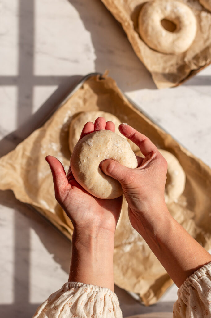 Two hands forming a bagel shape from dough with a baking tray of shaped dough balls in the background.