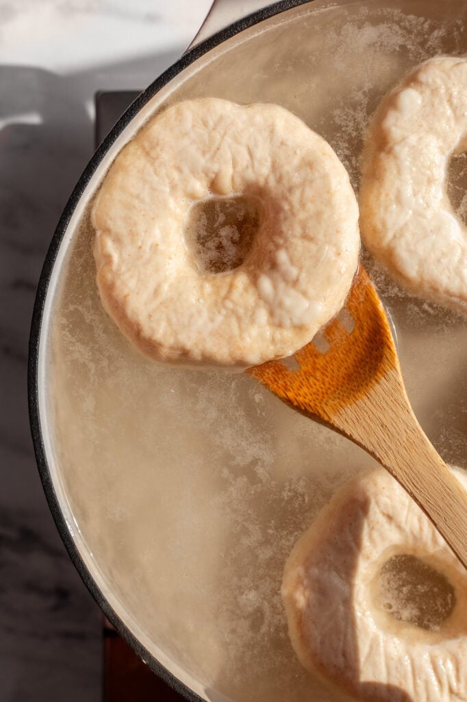 A wooden spoon lifting a freshly boiled sourdough bagel from a pot of bubbling water.