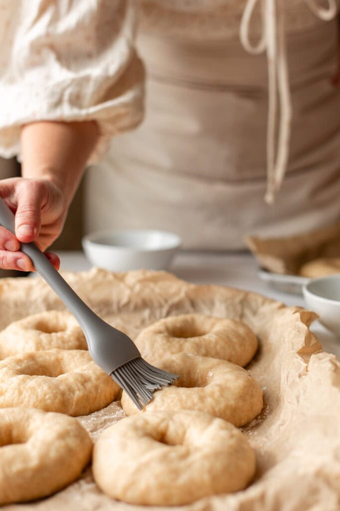 Brushing boiled bagels with egg wash on a parchment-lined tray before baking.