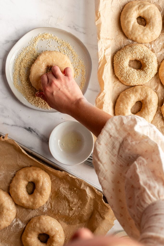 Hand dipping a boiled bagel into a plate of sesame seeds to coat the surface evenly.