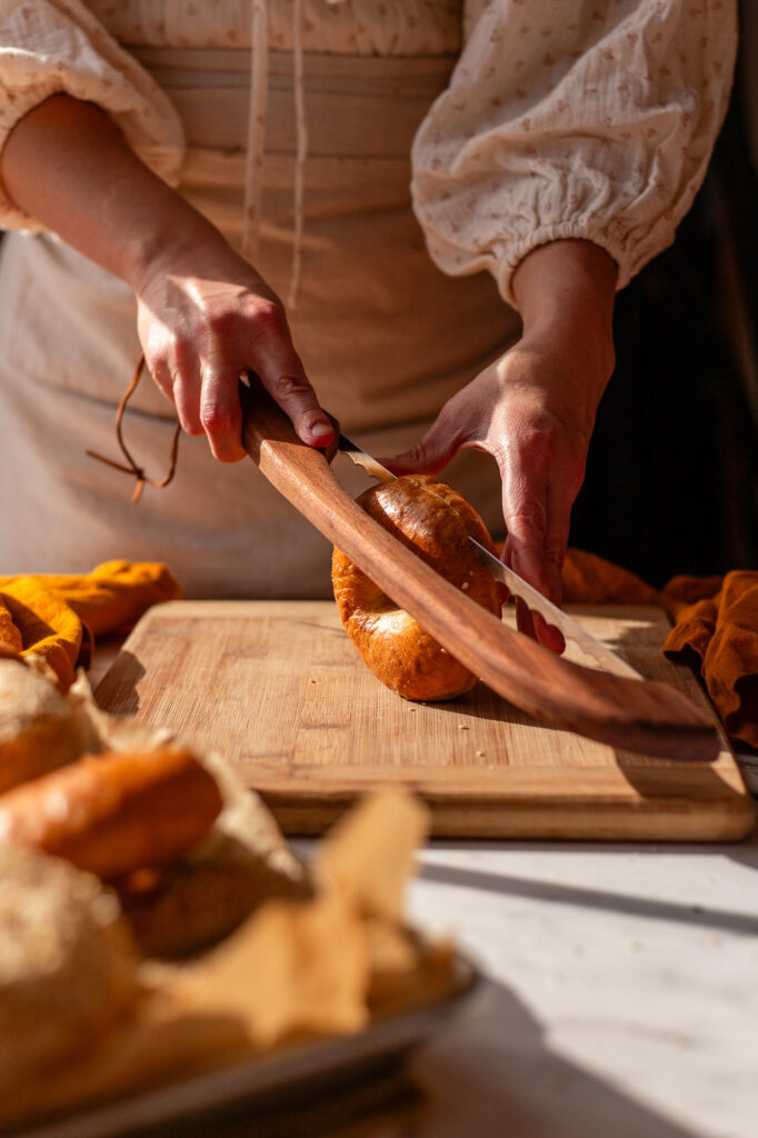 Hands slicing a freshly baked sourdough bagel with a serrated knife on a wooden cutting board.