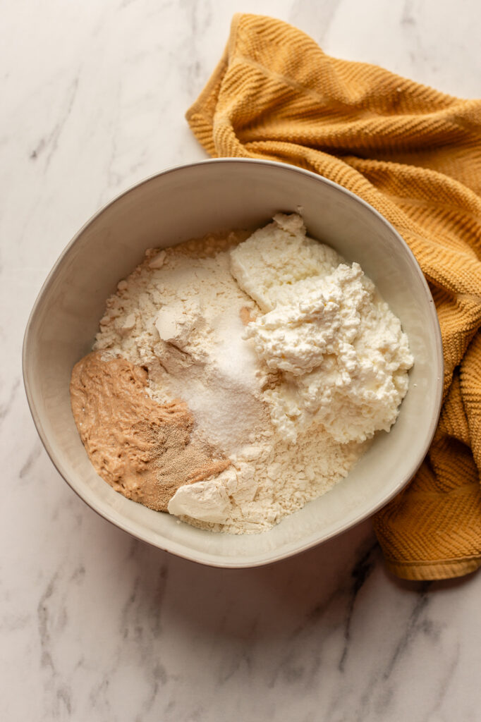 Ingredients for sourdough cottage cheese bagels in a mixing bowl, including flour, cottage cheese, sourdough starter, and yeast.