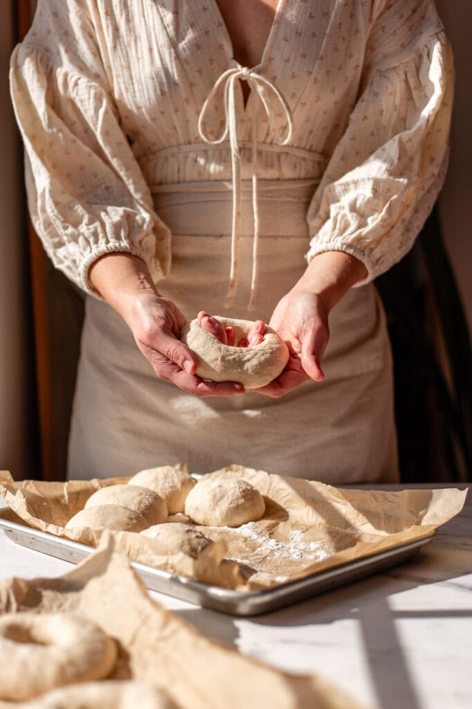 Hands shaping a ball of dough into a bagel by creating a hole in the center.