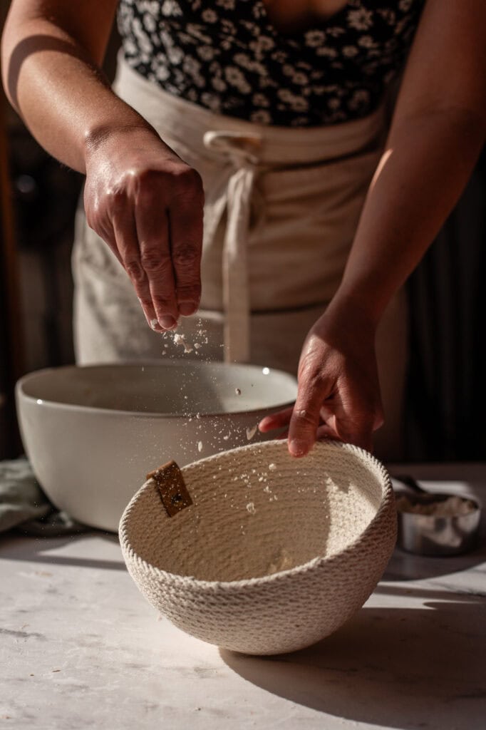 Sprinkling flour into a proofing basket in preparation for the final dough rise.