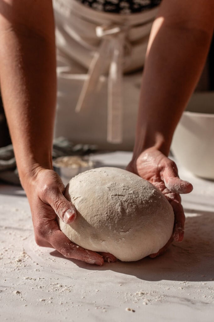 Hands cupping the dough to gently shape it into a smooth, rounded boule.