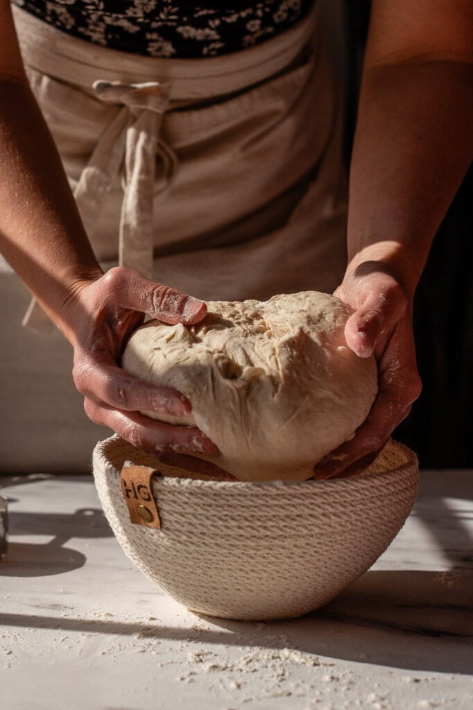 Hands carefully transferring a shaped sourdough boule into a floured proofing basket for its final rise.