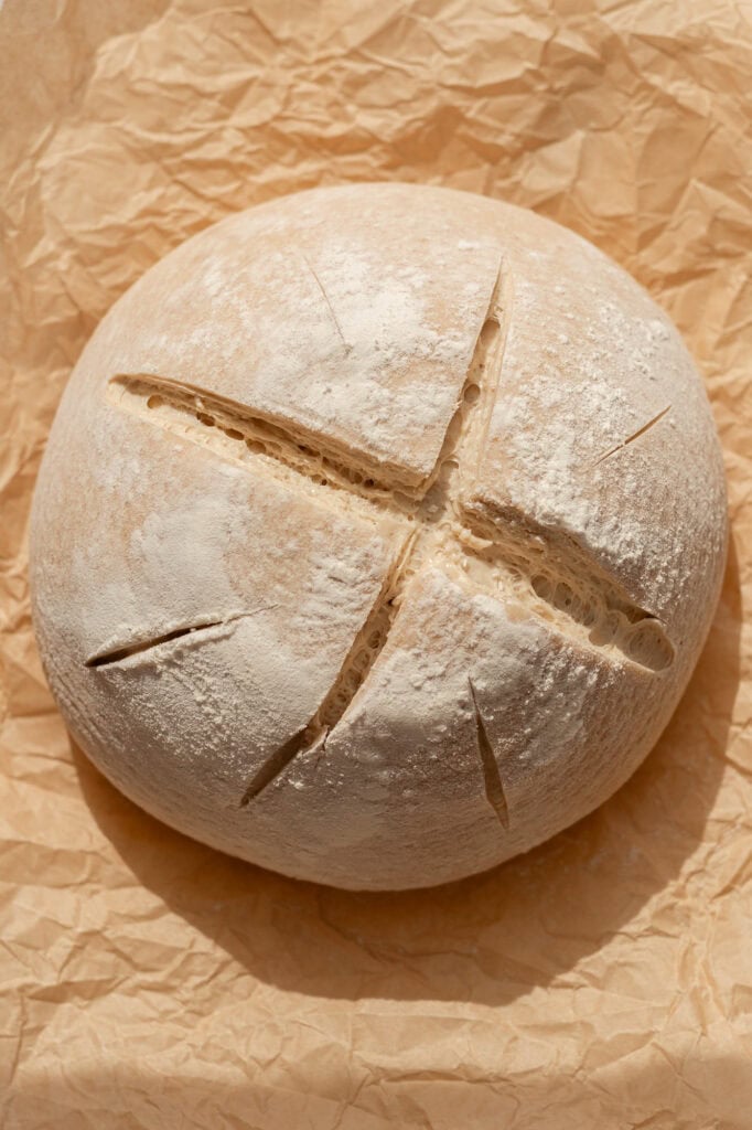 A scored sourdough boule dusted with flour, ready to be baked, resting on parchment paper.