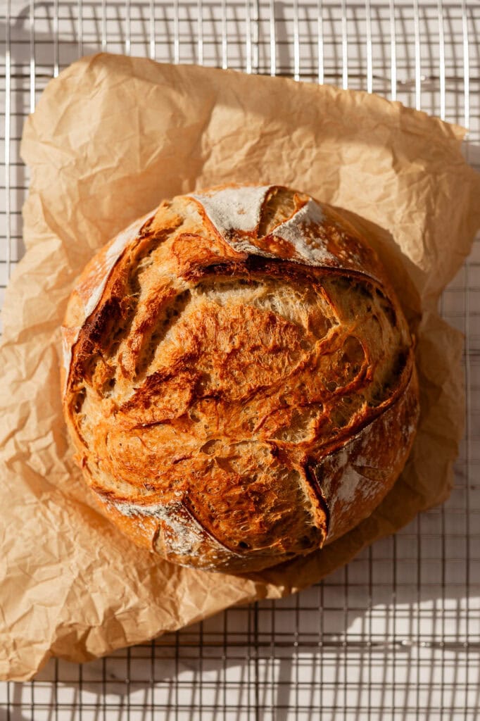 A fully baked sourdough boule with a golden crust, cooling on a wire rack lined with parchment paper.